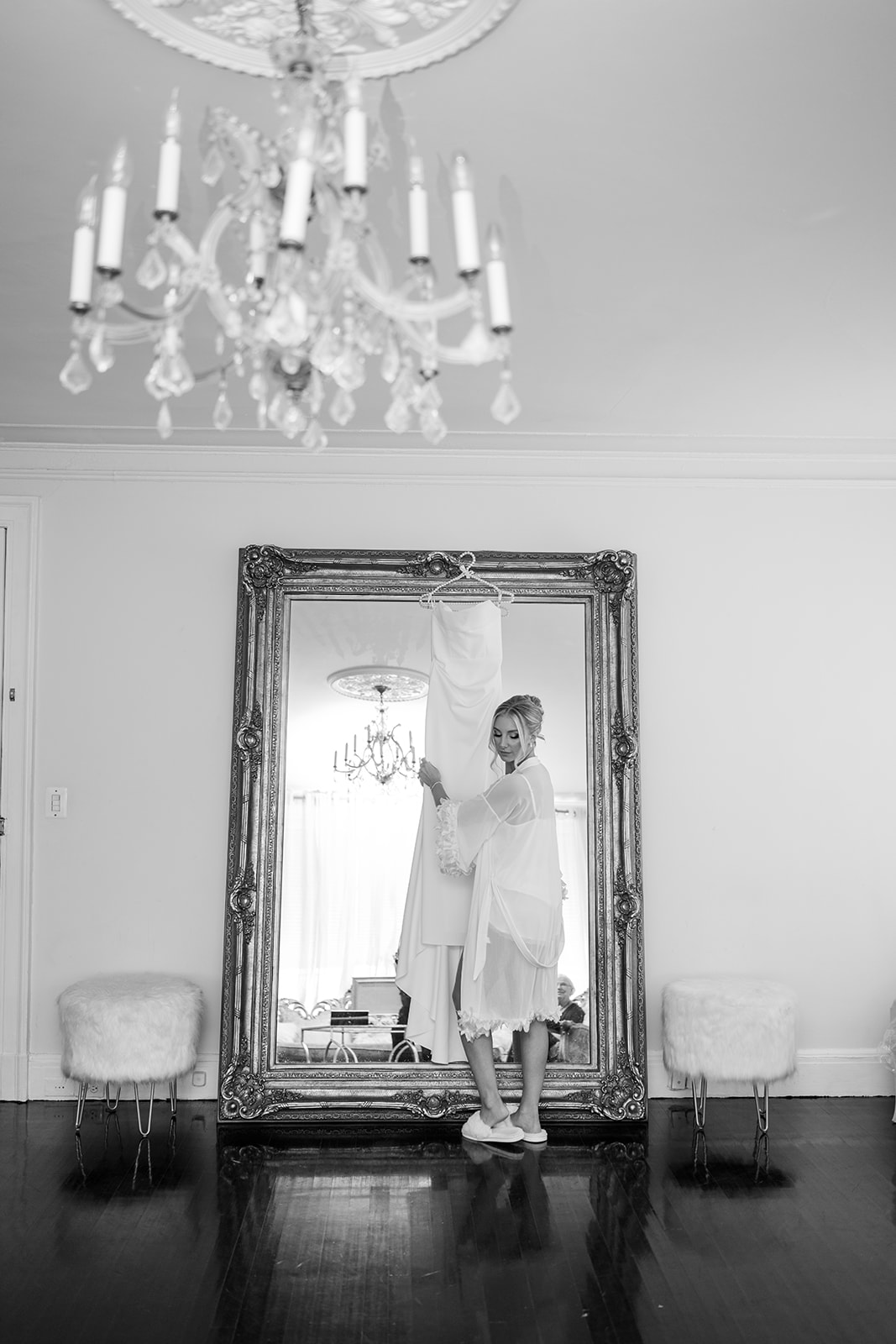 Black and white photo of the bride admiring her wedding gown in front of a grand, ornate mirror.